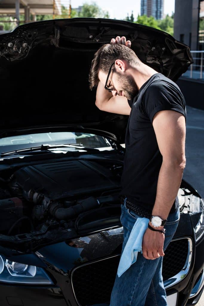 side view of confused man scratching head near broken car engine at street